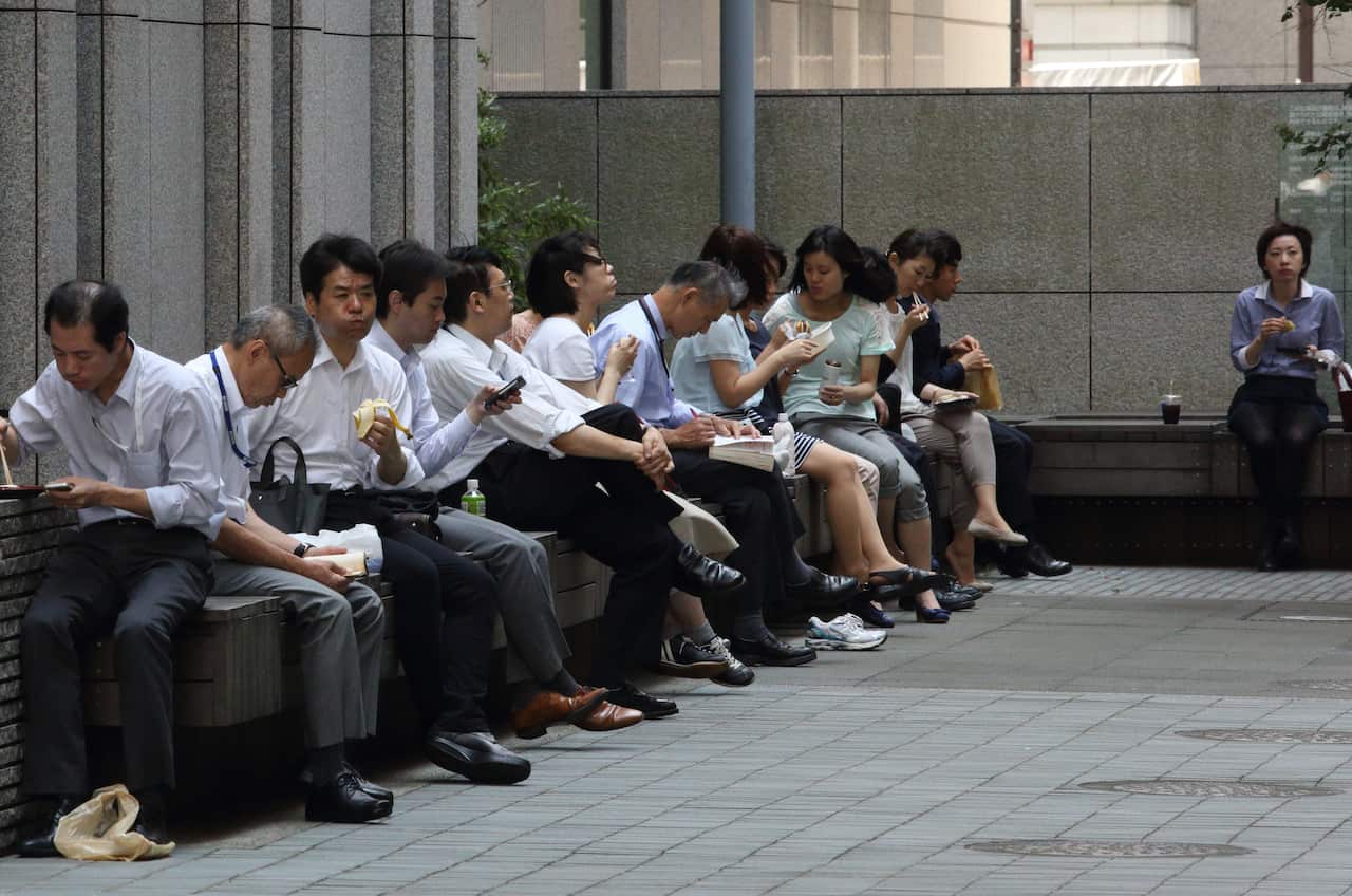 Workers in Japan sit on a bench eating their lunch.