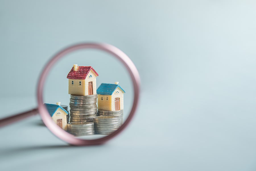 A magnifying glass is placed over three small toy houses sitting on piles of coins