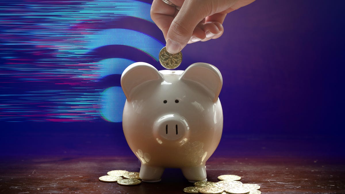 a hand depositing a coin into a piggy bank sitting on a wood table with coins scattered and a digitized wi-fi symbol in the background.