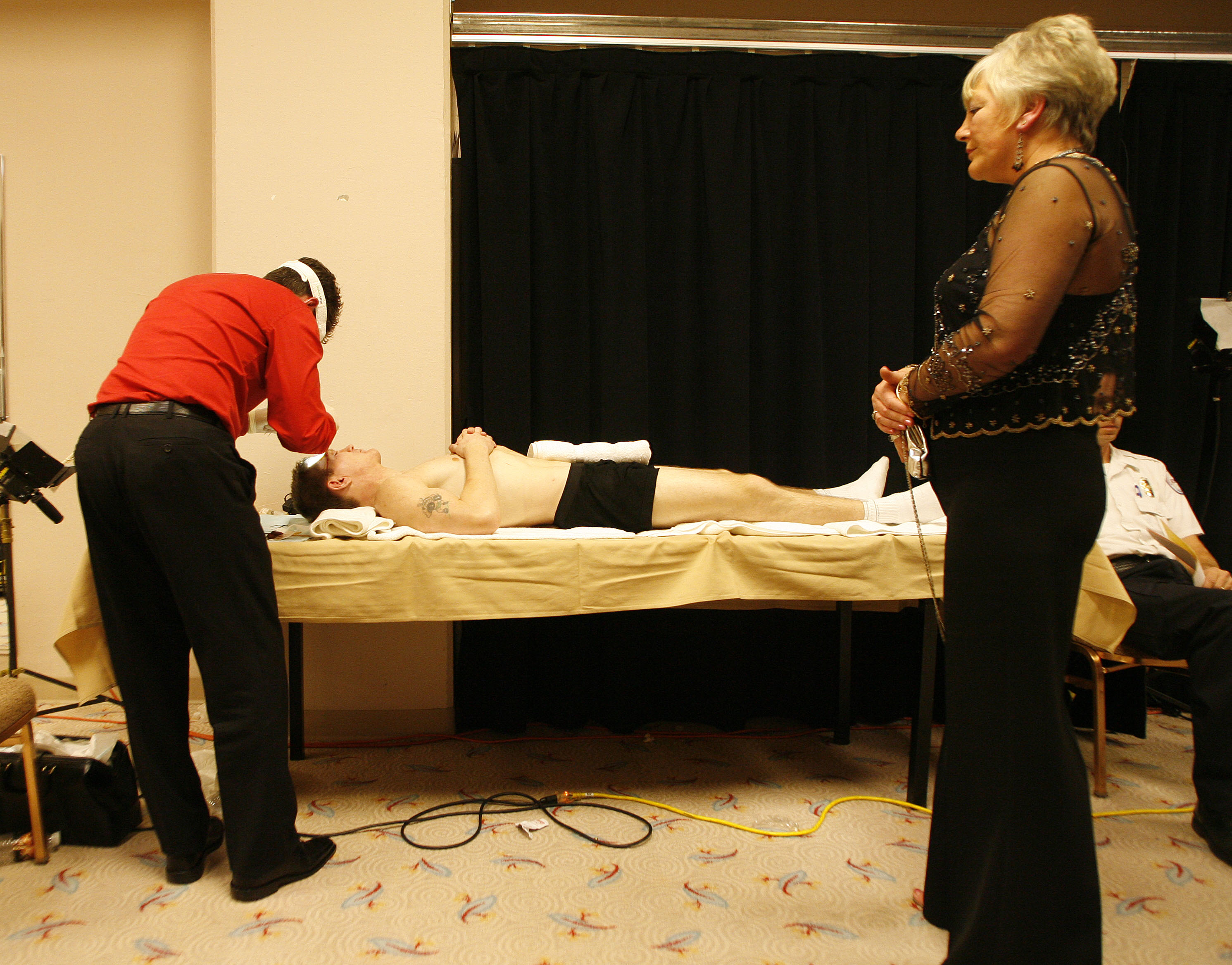 A man lying on a table while a person in a red shirt attends to him; a woman in a black dress watches.