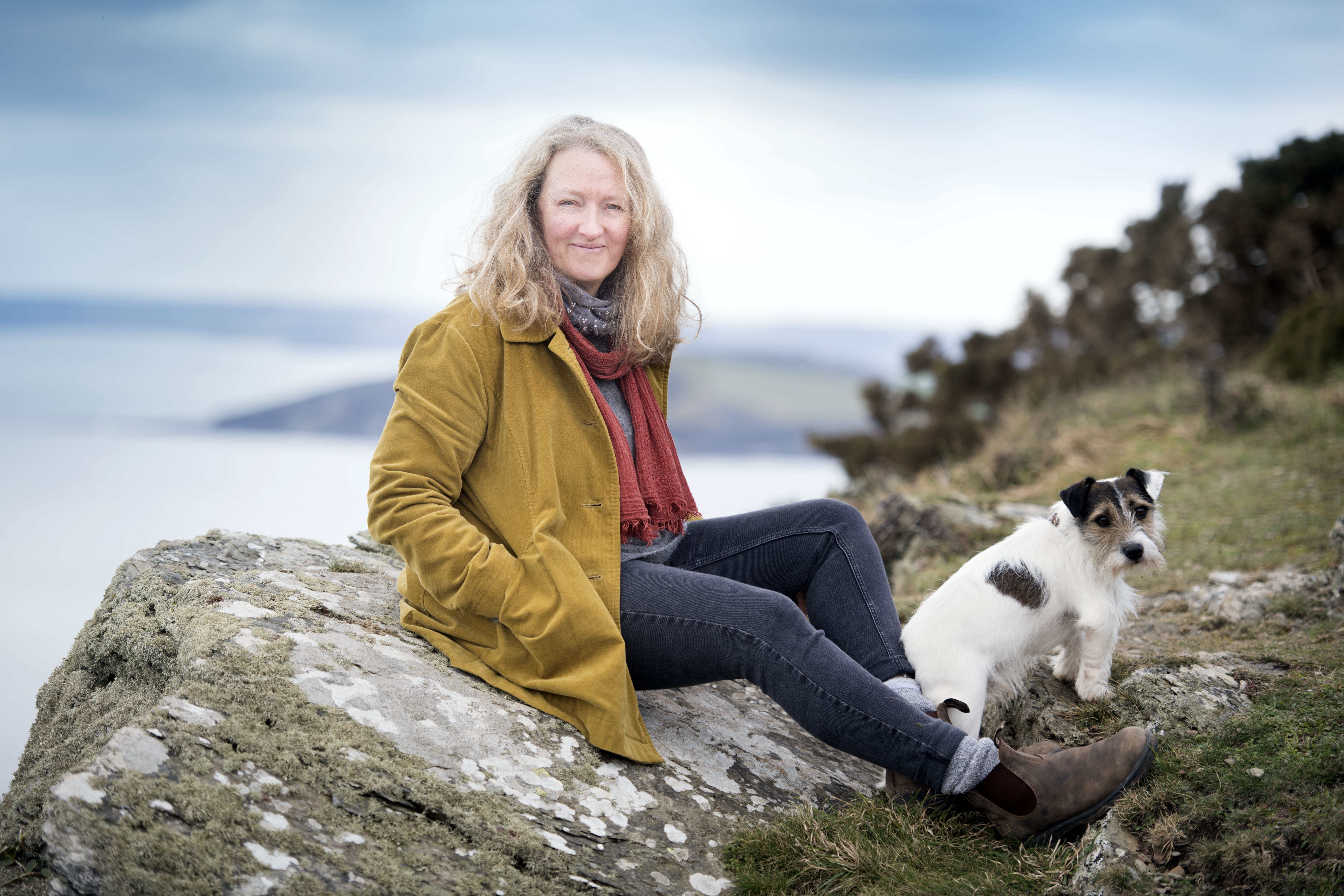 Raynor Winn sitting on a rock with her dog, overlooking the coast.