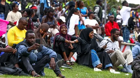Donwilson Odhiambo/Getty Images Football fans sitting outside on a grassy area. Some are smiling and others on their phone - Sunday 17 August 2025.