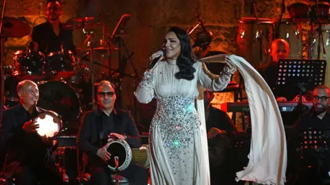 Fethi Belaid/AFP/Getty Images Ahlam performs on stage while wearing a white dress with jewels on it. Behind her is a band, including men in black suits playing a drum, tambourine and drums - Thursday 21 August 2025.