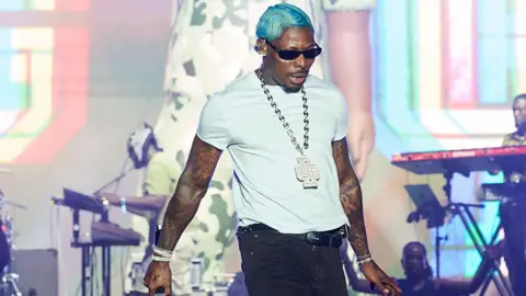 Robert Okine/Getty Images Singer Asake on stage with dark sunglasses wearing a white T-shirt and platinum jewellery. His band is behind him - Saturday 16 August 2025.