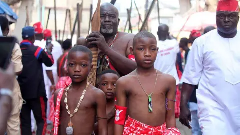Adekunle Ajayi/NurPhoto/Getty Images Young boys wearing red and white patterned traditional Yoruba clothes stand in front of men carrying what looks like sticks. Some of the men behind are wearing red and white clothing also - Wednesday 20 August 2025.