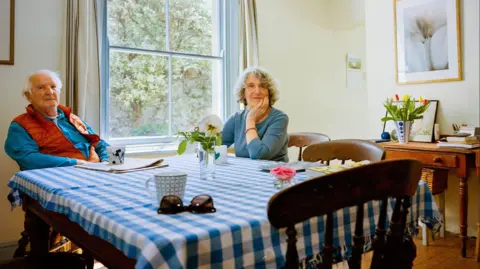 Mana Baoosh A man and a woman sit at a table covered in a blue gingham cloth. There are three mugs and a vase of flower sat on top and they are both smiling at the camera. In the background is a large window through which trees are visible