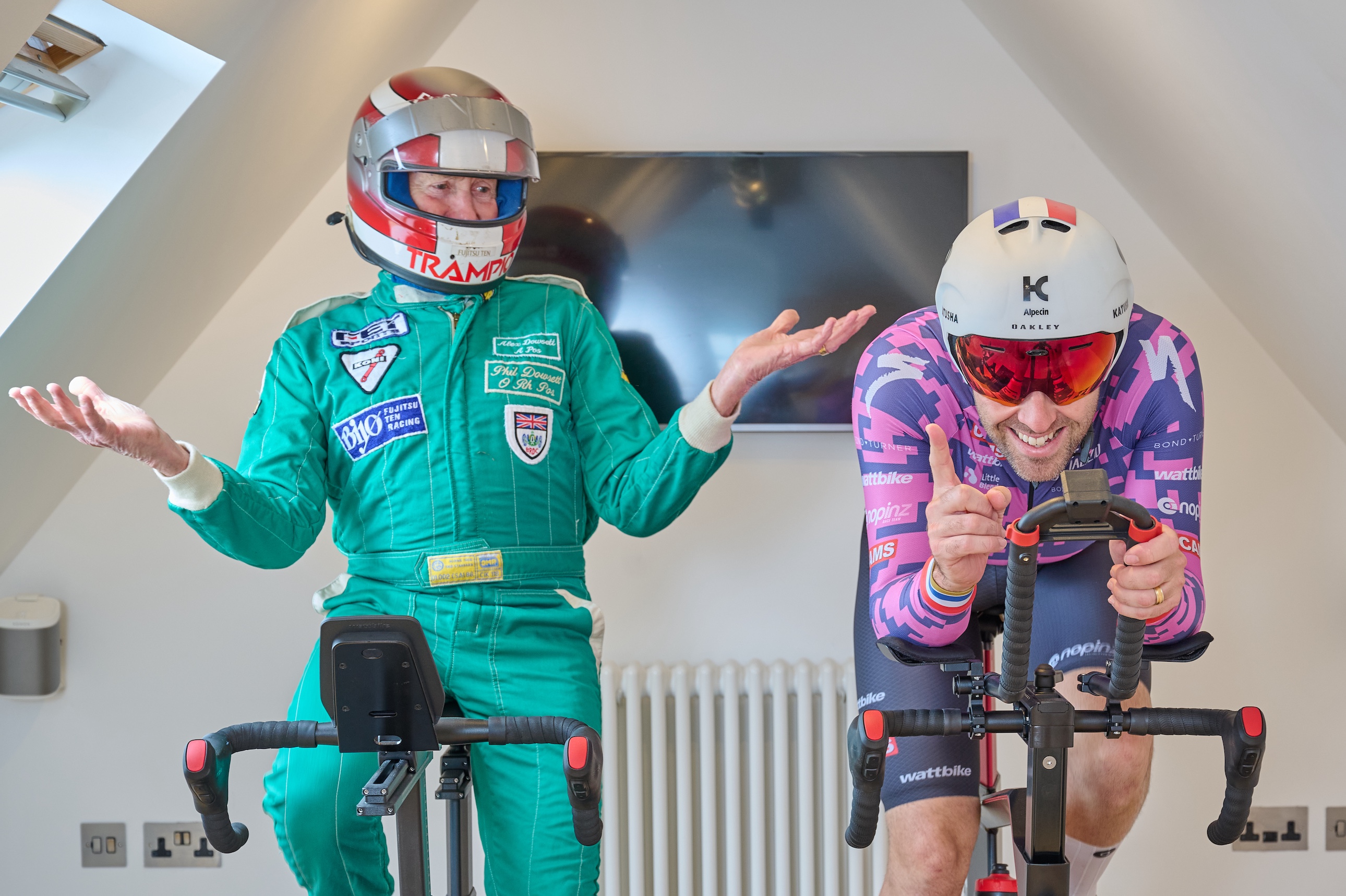 Alex Dowsett and his dad alongside each other on smart-bikes. Alex is wearing an aero helmet, his dad is wearing a full-face rally helmet