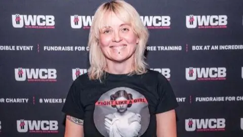 Teresa Colman Teresa wearing a black T-shirt - standing in front a UWCB poster - smiling for camera.