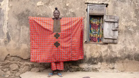 Emmanuel Adegboye/Shutterstock/EPA A woman wearing a red-checked traditional outfit spreads her arms to display her agbada style outfit. She is standing in front of a dishevelled brown coloured building - Saturday 16 August 2025.