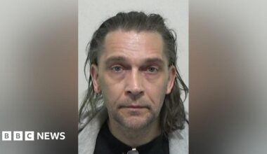 Mugshot of Taylor. They have very short stubble on their chin and upper lip and have shoulder length wavy brown hair