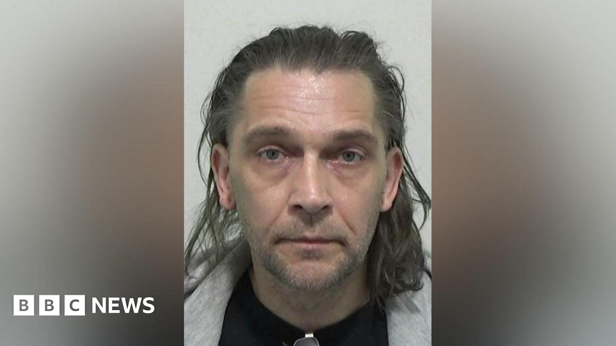 Mugshot of Taylor. They have very short stubble on their chin and upper lip and have shoulder length wavy brown hair