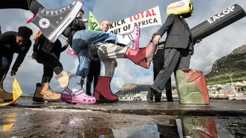 Brenton Geach/Gallo Images/Getty Images Protesters walking along the beach carrying Extinction Rebellion flags and a banner with the following words written on it "Kick Total Out Of Africa". The protesters are making kicking motions and have large mock shoes on - Tuesday 19 August 2025.