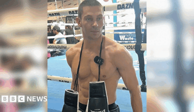 A man standing in front of a boxing ring, which has a blue floor. He is topless with a pair of black and white boxing gloves hanging around his neck. The image cuts off at the waistband of his black pair of shorts.
