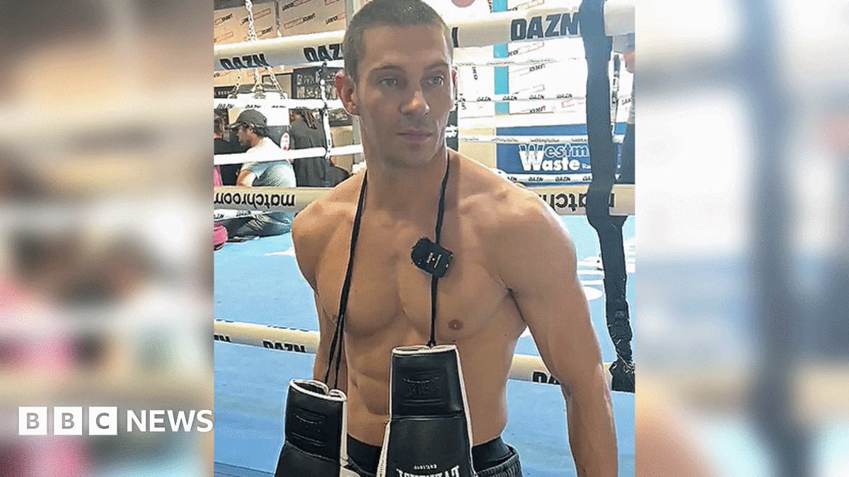 A man standing in front of a boxing ring, which has a blue floor. He is topless with a pair of black and white boxing gloves hanging around his neck. The image cuts off at the waistband of his black pair of shorts.