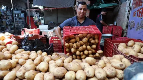 Billel Bensalem/APP/NurPhoto/Getty Images A man in a blue T-shirt tipping a red crate of potatoes into his market display. There is a large pile of potatoes in front of him - Sunday 17 August 2025.