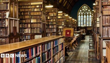 Ancient looking wood-panelled library with rows and rows of books and stained glass window in background.