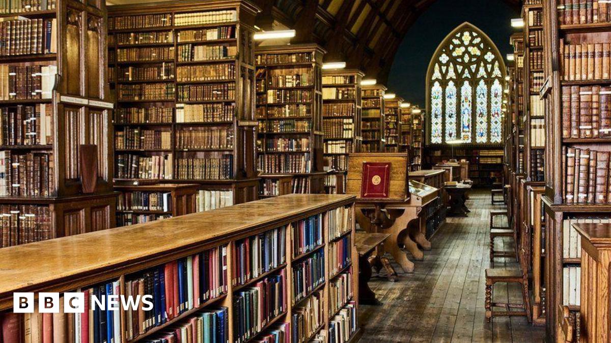 Ancient looking wood-panelled library with rows and rows of books and stained glass window in background.