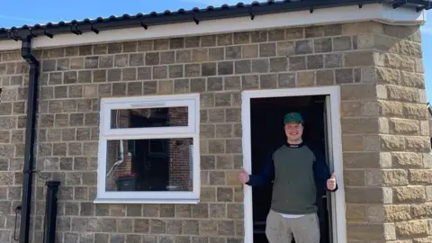 Chilli Charva A man stood in the doorway of what appears to be a very small brick house. It is actually an industrial kitchen, which he has built.