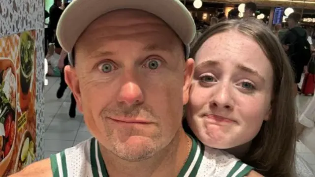 A man in a cap and white and green vest pulls a concerned face in a selfie, a younger girl with brunette hair is behind him