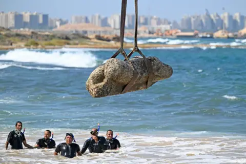 Khaled Desouki/AFP/Getty Images Divers in wet suits watch as a crane pulls an artefact from the waters at Abu Qir bay in Alexandria - Thursday 21 August 2025.