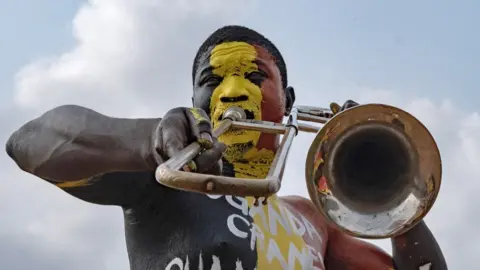 Badru Katumba/AFP/Getty Images A man blows his trombone against a background of blue skies, whilst wearing red, yellow and white face and body paint - Monday 18 August 2025.