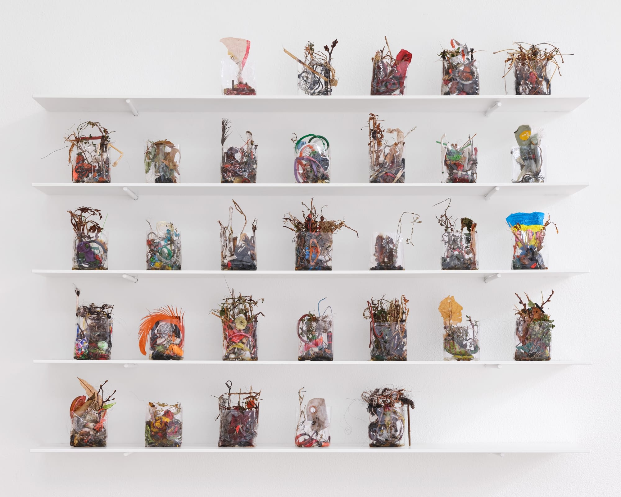 a collection of small ikebana-like sculptures in cellophane arranged like a month calendar on white shelves