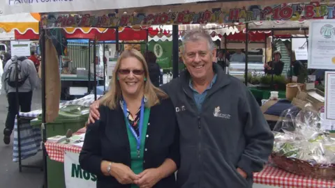 Moseley Farmers' Market A man stands by a market stall with his arm around a woman stood next to him. The man has short grey hair and is wearing a grey fleece. The woman next to him has long blonde hair and is wearing sunglasses with a dark jacket and a green top underneath. 