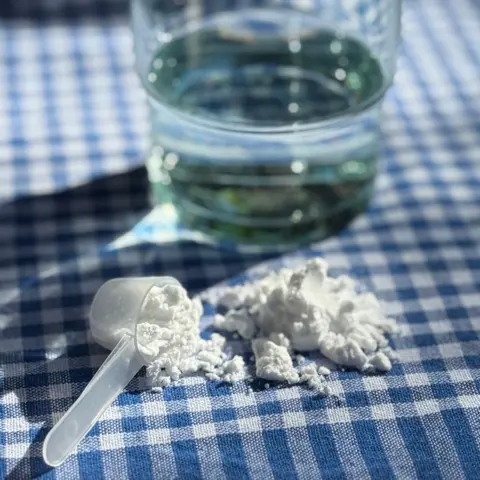 BBC Creatine powder and a scoop next to a glass of water on a blue and white check cloth