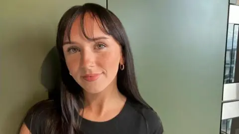 BBC Leoni Clancey, 21, with shoulder length straight dark brown hair with a wispy fringe wearing a black t shirt standing against a khaki wall with a neutral expression
