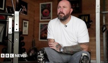 Dave Watson is sitting on a bench in his gym at home. He is wearing a white t-shirt and grey shorts. He has two dark black prosthetic legs covered in stickers. Behind him on the wall are photographs including one of him winning shootout gold medal at the Invictus Games in Sydney.