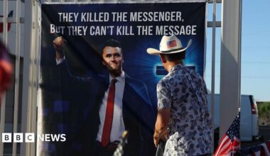 A man wearing a cowboy hat with a US flag on the back looks at a banner with a large picture of Charlie Kirk on it - the message on the banner reads "they killed the messenger, but they can't kill the message"