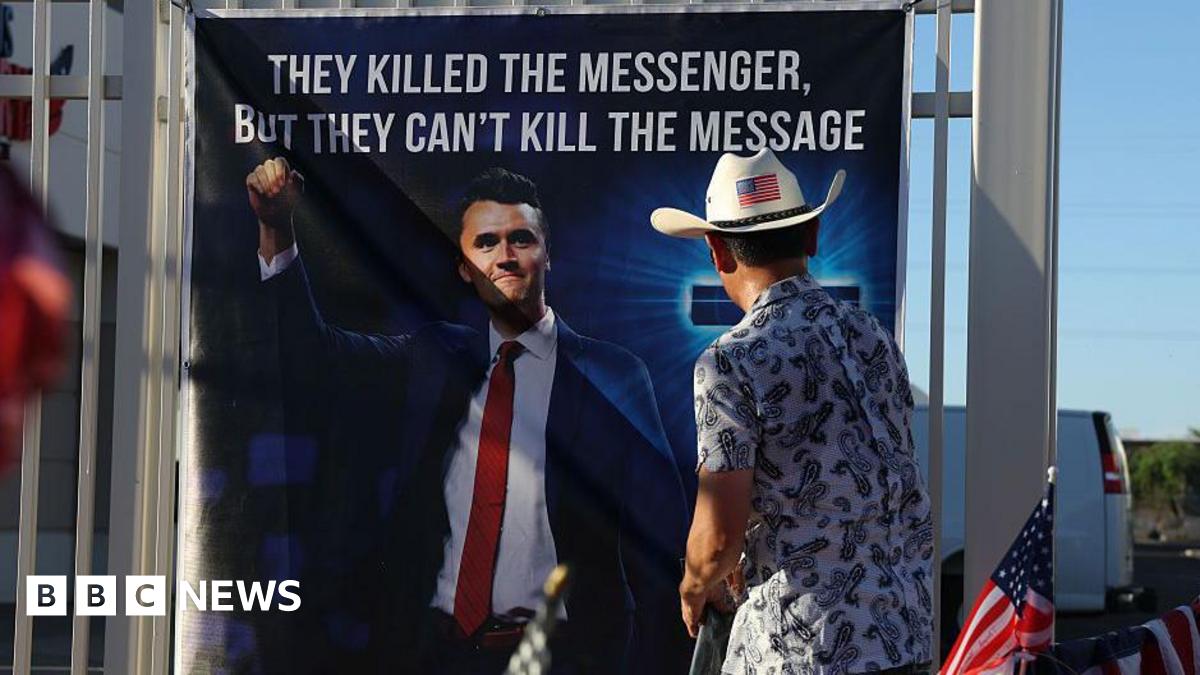 A man wearing a cowboy hat with a US flag on the back looks at a banner with a large picture of Charlie Kirk on it - the message on the banner reads "they killed the messenger, but they can't kill the message"