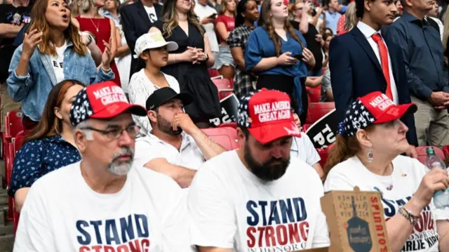 Three people sit in the stadium stands wearing hats reading "Make America Great Again" - others stand behind them