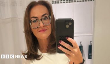 A woman with straight brown hair, a white t-shirt, patterned glasses and pink lipstick takes a selfie in the mirror with a phone