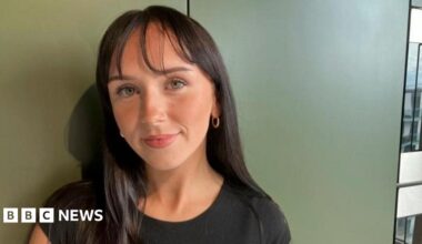 Leoni Clancey, 21, with shoulder length straight dark brown hair with a wispy fringe wearing a black t shirt standing against a khaki wall with a neutral expression