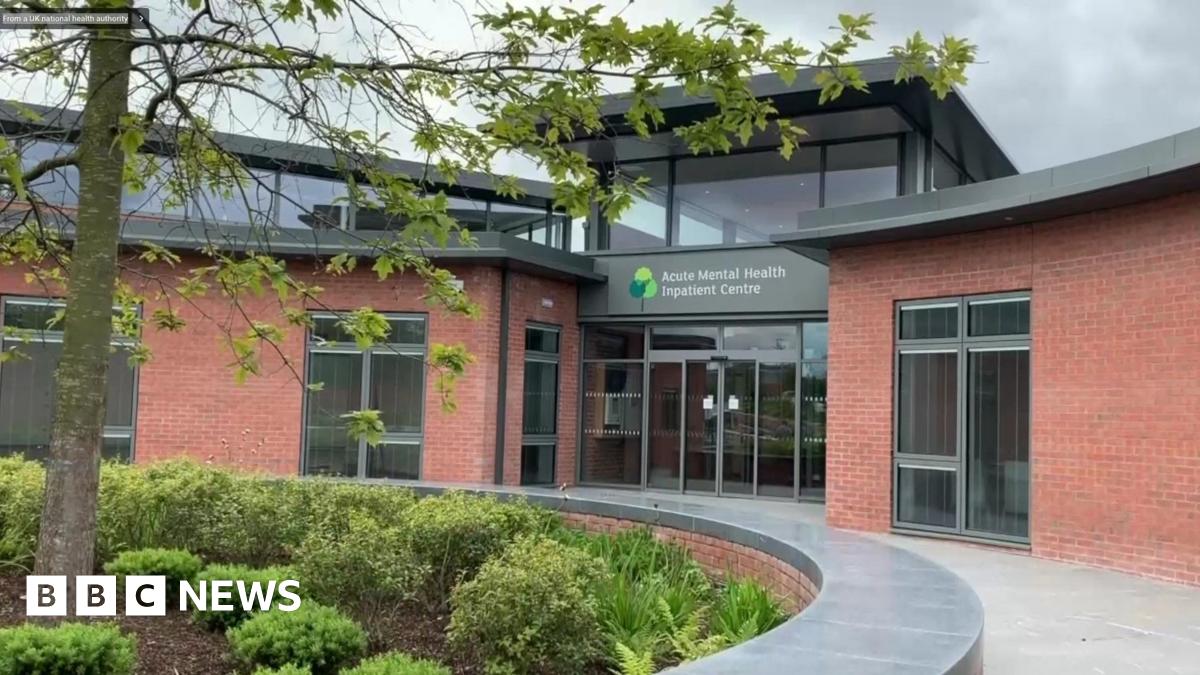 Green signage reading 'Acute Mental Health Inpatient Centre'. The sign is in front of a red brick wall and it is partially covered by a bush.