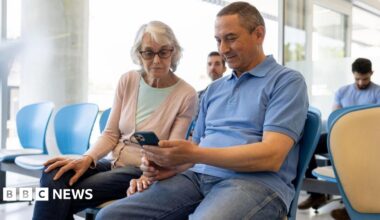 A waiting area with several people seated on blue and beige chairs. In the foreground, a man and a woman are sitting together and looking at a smartphone.