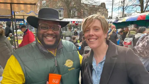 Moseley Farmers' Market A man wearing a black hat, glasses and a green gilet is stood with a woman with short blonde hair, who is wearing a brown coat with a denim top. 
