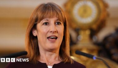 A woman with shoulder-length brown hair and a fringe speaks into a microphone, with a gold clock featuring prominently behind her. She wears a black cardigan and a gold necklace, with small silver hoop earrings.