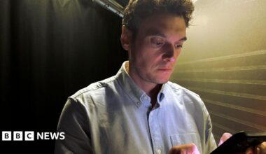Joe Tidy looking down at a phone. He has short brown hair and is wearing a light blue shirt.