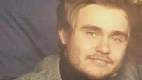 Jesse Lewis Jac Lewis smiles at the camera, he has long sandy hair and is wearing a grey blazer.