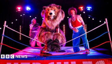 A theatrical scene set in a wrestling ring. A performer in a bear costume stands centre stage, flanked by two other characters in colourful outfits. One wears blue pants and a red tank top, striking a confident pose.