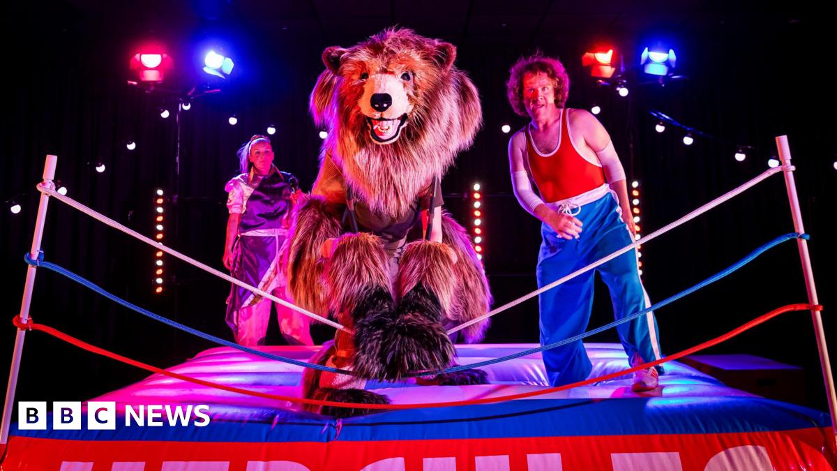 A theatrical scene set in a wrestling ring. A performer in a bear costume stands centre stage, flanked by two other characters in colourful outfits. One wears blue pants and a red tank top, striking a confident pose.