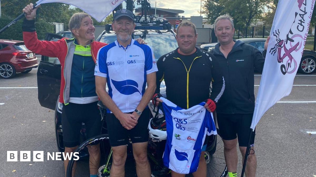 Manfred Saenger, Tim O'Leary, Russell Smith and Patrick Brewster, standing outside, in front of a bike. Manfred is looking to the side and holding up a flag; Tim is wearing black shorts and a white and blue top, a cap and is smiling at the camera; Russell is wearing black, holding up a cycling top; and Patrick is also holding a flag.