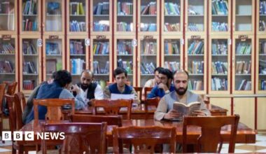 Men sit at tables in a room with bookshelves in the background