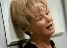 Person with short blond hair, leaning against a wall, appears distressed or emotional, mouth open as if crying or speaking