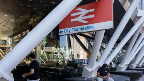Getty Images A railway station with a large sign bearing the railway symbol and the words City Thameslink. There are white roof support poles in the foreground, and a Starbucks Coffee in the background. A man is sitting below the sign looking at his mobile phone.