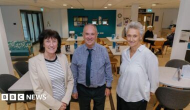Becky Land holding a bucket in her left hand. Her right hand is placed on a black and white cow which is looking at the camera. There are more cows in the barn behind.