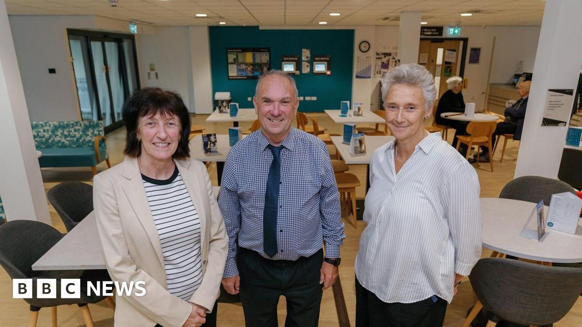 Becky Land holding a bucket in her left hand. Her right hand is placed on a black and white cow which is looking at the camera. There are more cows in the barn behind.