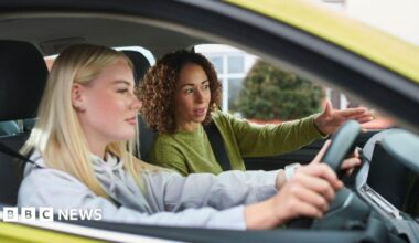 Two women in a car. The younger one is at the wheel. She is white and has blonde hair and is wearing a hoodie. An older mixed-race woman is instructing her. The car is yellow-green